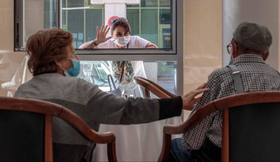 Un familiar saluda por la ventana a dos ancianos en una residencia de mayores.